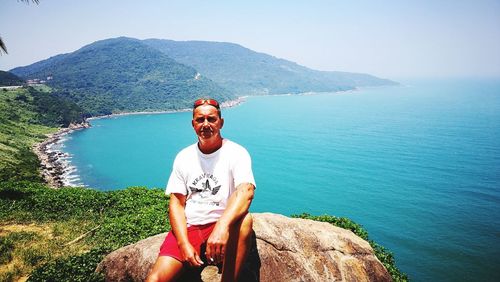 Portrait of young man sitting on sea against mountain