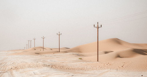 A continuous row of electricity pole erected in the desert with dunes in the background.