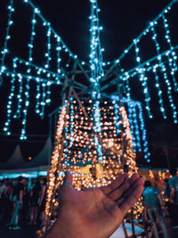 Cropped hand of person against illuminated christmas lights at night