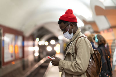 Black man waiting for train at subway station, using mobile phone, wear face mask. covid-19 pandemic