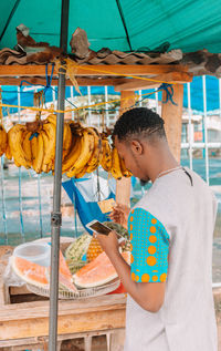 Rear view of man using mobile phone while standing by fruit stall