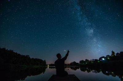 Silhouette man pointing at lakeshore against sky at night