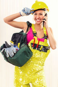 Portrait of female worker in coverall with tool bag talking on phone against wall