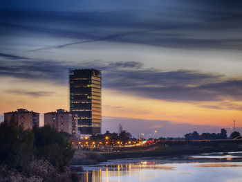 Illuminated buildings by river against sky at sunset