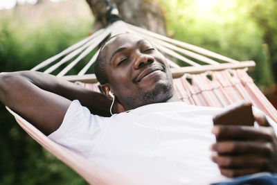Smiling man with closed eyes enjoying music through mobile phone while relaxing on hammock