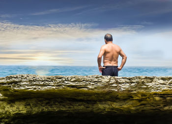 Rear view of shirtless man standing on rock by sea against sky