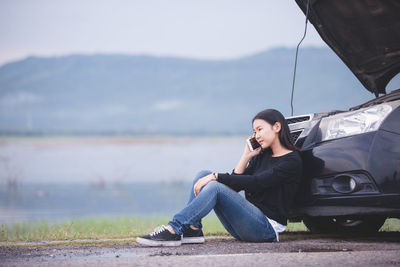 Young woman looking away while sitting on car against sky
