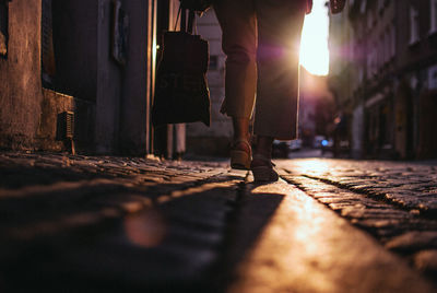 Low section of woman carrying shopping bag while walking on footpath during sunset