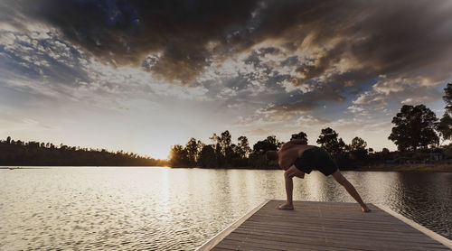 Shirtless man practicing yoga on pier against sky during sunset