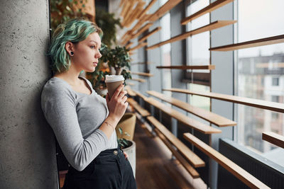 Alternative girl in casual clothes and with green hair standing with cup of drink in hands.