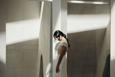 Side view of calm pensive young ethnic woman in elegant white wedding dress standing alone near column in room with sunlight and shadows