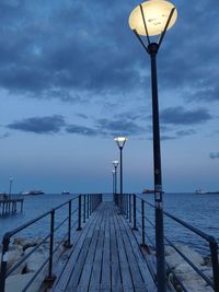 Street lights on pier by sea against sky at dusk