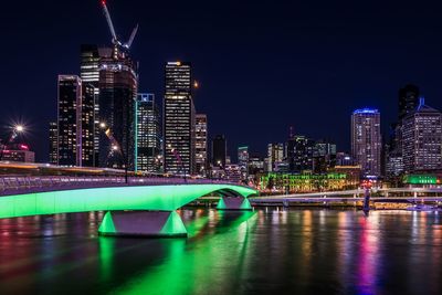 Illuminated bridge over river by buildings against sky at night