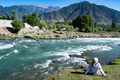 Rear view of woman sitting on rock by sea against mountain