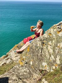 Woman having drink while sitting on rock formation by sea