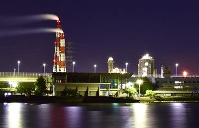 Illuminated factory by river against sky at night