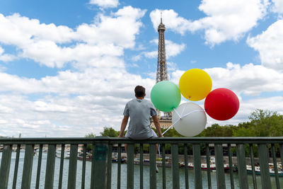 Rear view of man sitting with balloons on railing against eiffel tower