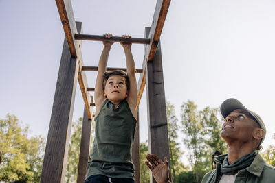 Male camp counselor assisting boy doing monkey bars at summer camp