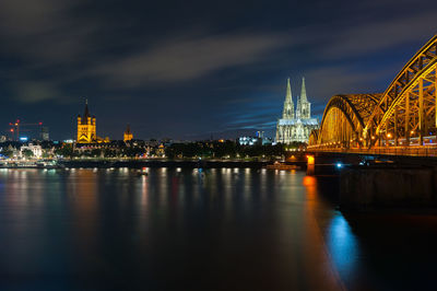 Illuminated bridge over river by buildings against sky at night