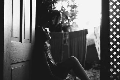 Side view of sad woman sitting by door against laundry drying in yard