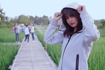 Portrait of mid adult woman wearing hooded shirt standing on boardwalk against sky