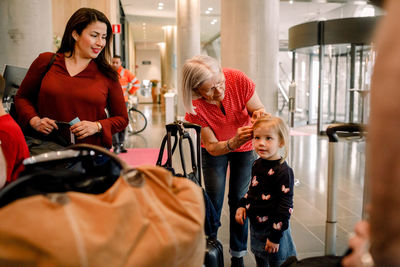 Smiling mother looking at senior woman tying daughter's hair while standing in hotel