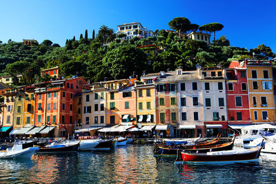 Boats moored in sea by buildings against blue sky