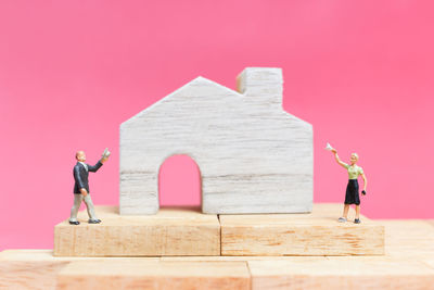 Close-up of figurines with model home on table against pink background