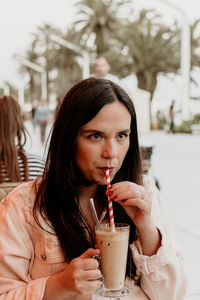 Young woman holding drinking straw in glass of iced coffee in bar in city