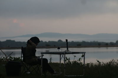 Men sitting on seat by lake against sky during sunset