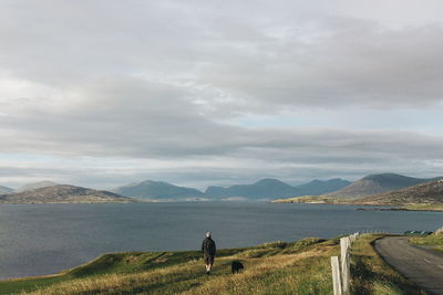 Rear view of man standing with dog on field by sea against cloudy sky