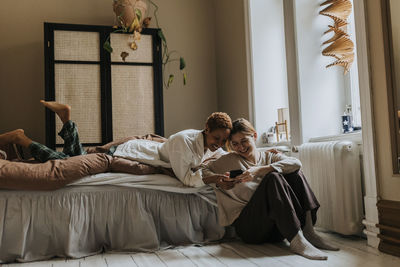 Smiling non-binary couple sharing smart phone while spending leisure time in bedroom at home