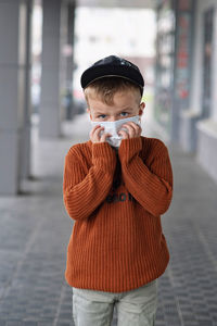 A boy in a cap and an orange sweater holds a medical mask with his hands.