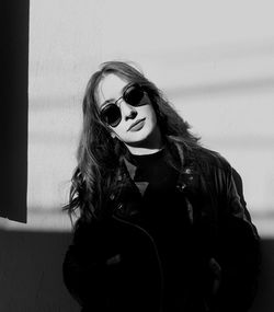 Portrait of young woman wearing sunglasses and jacket against wall