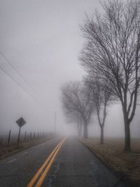 Road by bare trees against sky during foggy weather