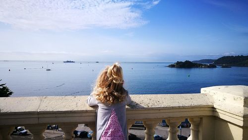 Rear view of girl looking at sea while standing by railing against sky