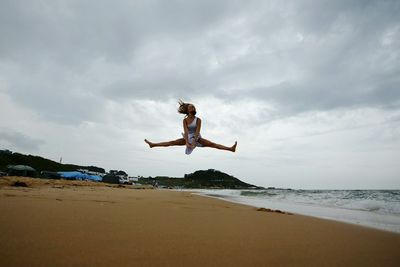 Low angle view of woman doing the splits over beach against cloudy sky