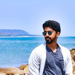 Young man wearing sunglasses standing at beach against clear sky