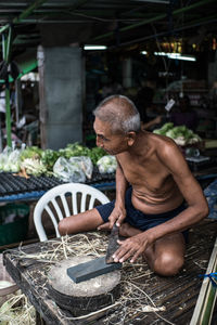 Shirtless senior man sharpening knife on seat at market