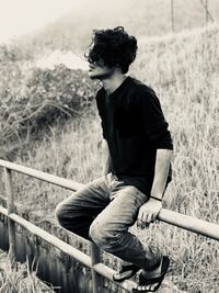 Full length of young man sitting on fence at field