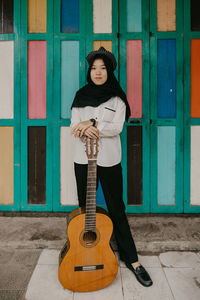 Full length portrait of woman holding guitar against wall