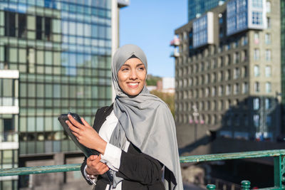 Cheerful muslim female entrepreneur in hijab and with takeaway coffee standing in street