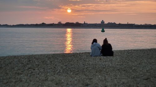 Rear view of couple sitting on beach against sky during sunset