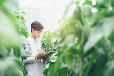 Young man analyzing plants with paper and pen while standing in farm