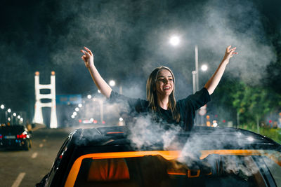 Happy young woman with arms raised through sun roof of a car in the city at night