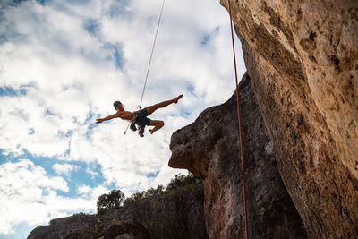 Low angle view of man hanging from safety harness by rock formation against sky