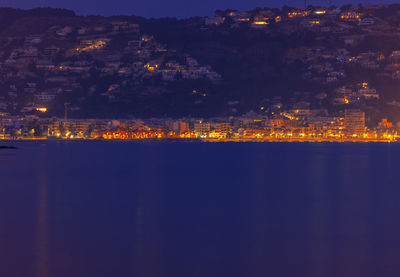 Illuminated buildings by sea against sky at night