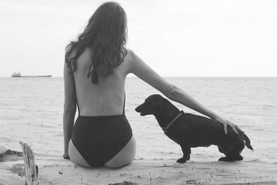 Rear view of woman in swimsuit sitting dog sitting on fallen tree trunk at beach against sky