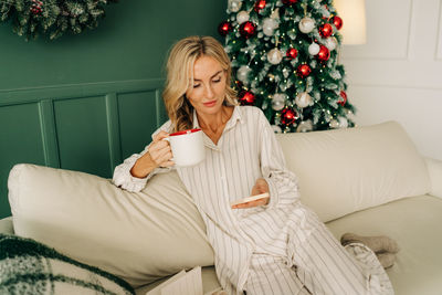 A woman sitting on a sofa in a christmas living room uses a mobile phone and drinks coffee.