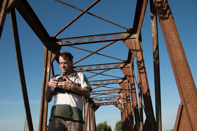 Low angle view of man holding camera standing on built structure against clear blue sky